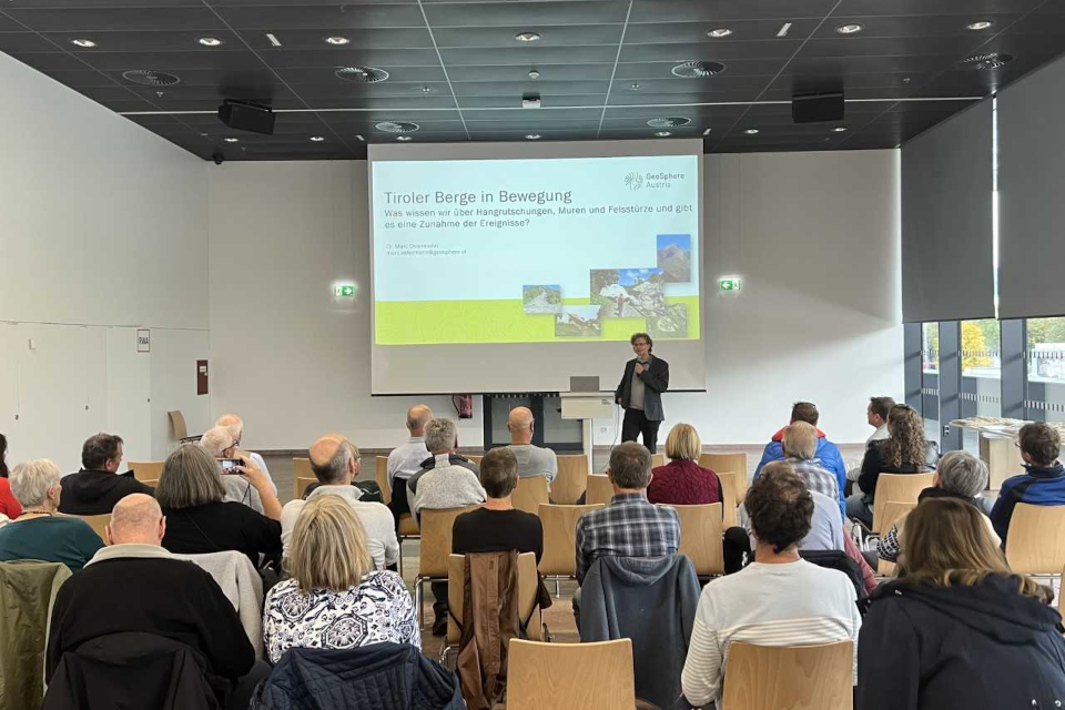 Group of people sitting in a room where a man is giving a lecture with a screen in the background 