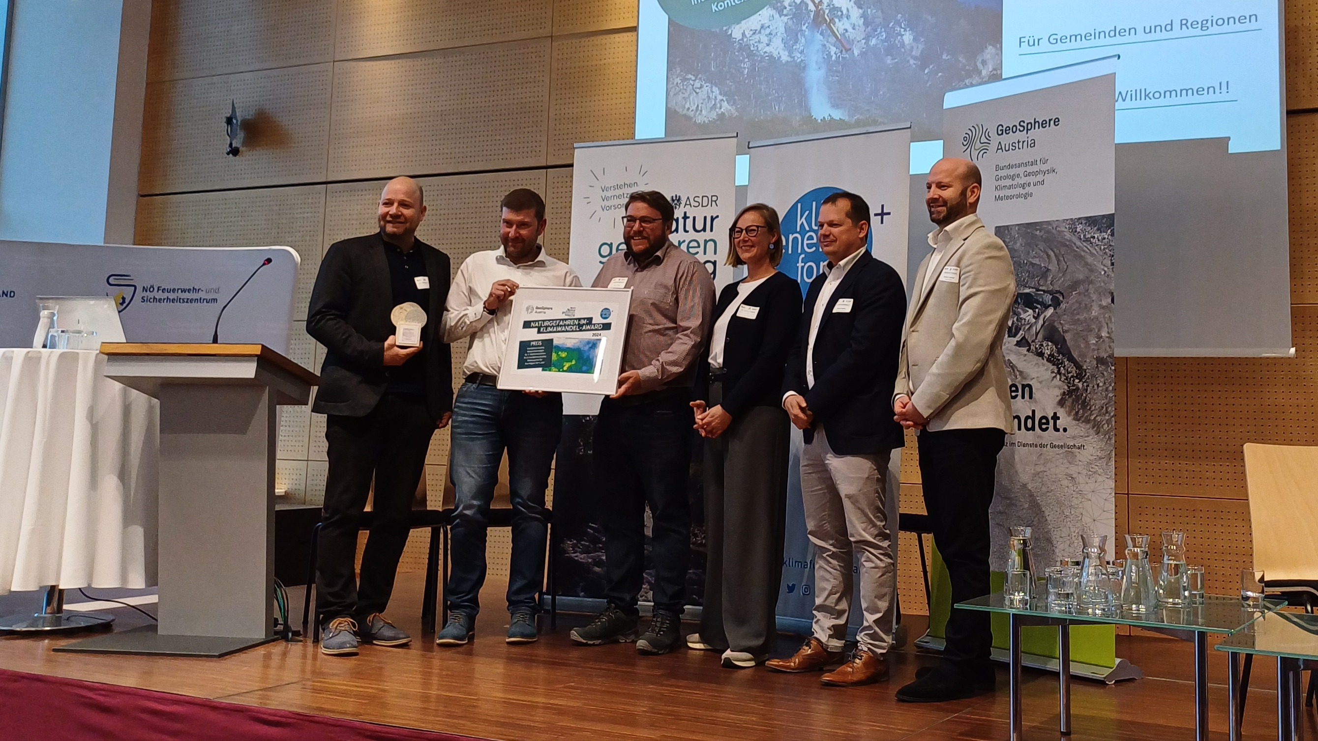 6 Personen stehen nebeneinander auf einem Podium und halten den Preis in Händen. Dahinter eine Leinwand mit einer Präsentationsfolie.